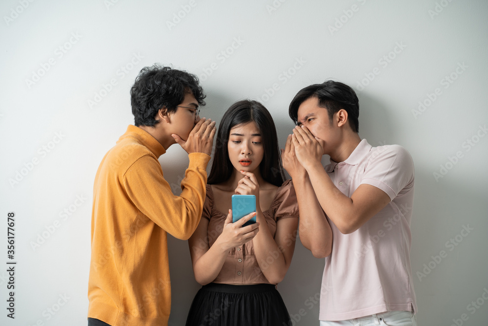 Wall mural two young men whispering to a young girl using a smart phone while standing against an isolated background