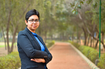 Closeup portrait of a confident young Indian Corporate professional woman with short hair and spectacles, crossed folded hands in an outdoor setting wearing a black business / formal suit