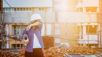 Working woman wipe off the sweat during survey construction site. Portrait of Female engineer holding Blueprints with the background of buildings and pile of bricks during construction.