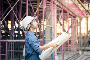 Asian woman architect or construction engineer wearing white hard helmet hold blueprint inside a construction site with scaffolding in the background. Engineer work concept.