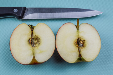 Two half of a red white apple and a kitchen knife on a light blue textured background close up