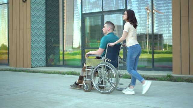 A young woman is carrying a young man in a wheelchair. An assistant carries a disabled person in a wheelchair along the street. Special transport for the disabled.