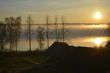 Leningradskaya oblast Russia, May 05 2016. Sunrise over the lake in the fog. Smoke over water and silhouettes of small houses and trees