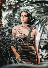 Fototapeta na wymiar Gorgeous young 20s woman wearing long evening elegant dress and tiara on head posing surrounded by lush tropical foliage trees. Beauty and fashion concept.