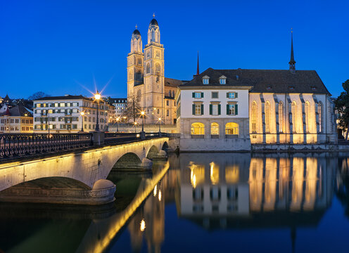 View of the illuminated Grossm&uuml;nster cathedral and Wasserkirche church across the river, reflecting on the water at dusk, at the old town Zurich, Switzerland