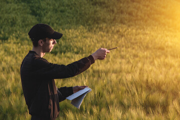 Young agronomist in cap takes notes in a notebook on a green agricultural field during sunset