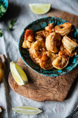 Turquoise bowl with steaming roasted garlic shrimps, lemon quarters, fresh baguette and bowl with greenery on linen tablecloths.