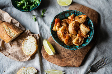 Turquoise bowl with steaming roasted garlic shrimps, lemon quarters, fresh baguette and bowl with greenery on linen tablecloths.