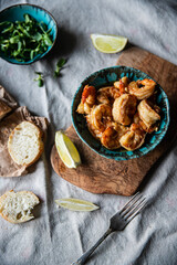 Turquoise bowl with steaming roasted garlic shrimps, lemon quarters, fresh baguette and bowl with greenery on linen tablecloths.