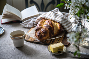 Homemade braided bread (challah) on wooden board half covered with a towel, butter, cup of coffee...