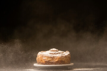Icing sugar is sprinkling over fresh bundt cake. Freezing motion. Copy space, black background. Bakery concept. Sweet bread on a white plate on a black background.
