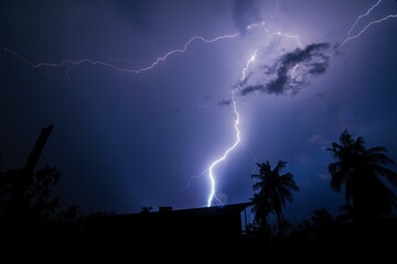 Thunderstorm strikes through the sky making bright light. Silhouette of homes and trees underneath