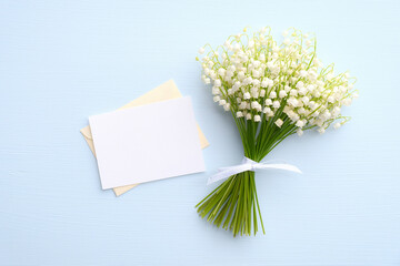 Bouquet of spring flowers lily of the valley and romantic letter with blank paper card on blue background. Flat lay, top view. Mothers Day, Women's Day, wedding greeting card concept.