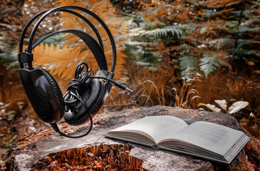 Black headphones and a white book in the forest on a stump against a background of green and orange...