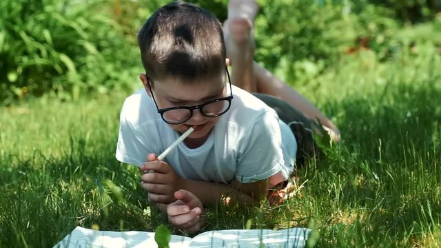 portrait of school boy in glasses studying outdoors on grass at school garden. male kid writing drawing in copy book in natural. little child thinking over homework. focused smart preschool child