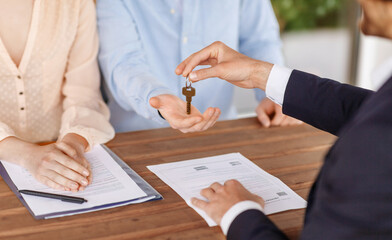 Real estate agent giving house key to young couple after signing property agreement at table, close up