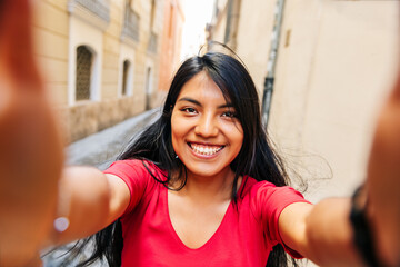 Close-up selfie of brunette Latin girl smiling holding the camera with both hands, on a European city street.