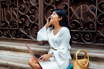 Brunette Latin woman with long hair sitting on stone staircase with laptop and talking on the phone while smiling