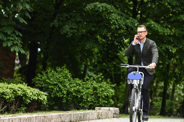 Use environmentally friendly modes of transport. Cheerful young man in smart casual wear and eyeglasses is riding a bike and talking on the phone while getting to the office