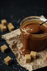 Glass jar of homemade salted caramel with a spoon and cubes of cane sugar