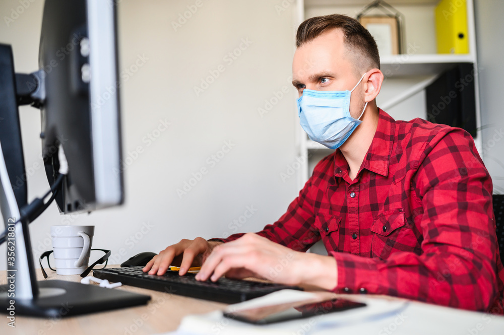 Wall mural responsible employee works with the computer wearing a protective mask. a young man works in the off