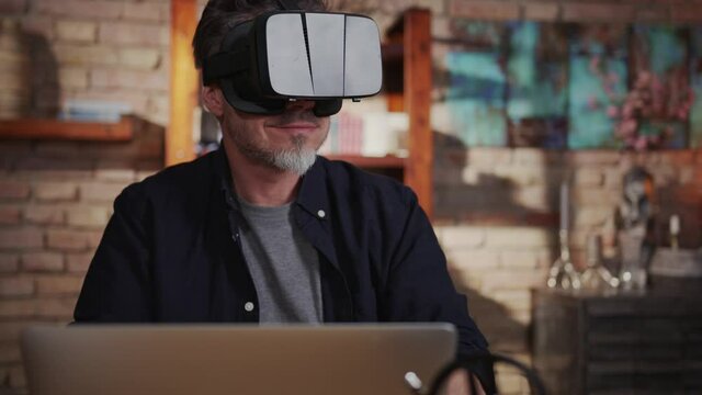 Casual man man working with virtual reality googles at home sitting at desk with laptop computer in home office.