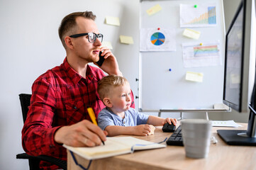 A young man works from home while quarantine, he sits at the computer and talks on the phone, his kid sits on his lap. Young dad works in home office with a kid in front of a PC