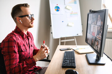 Online meeting of a young man with his subordinates. He sits accross from the window talking via web camera on PC to a group of people at the workplace, flipchart on the background