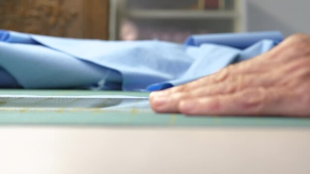 Side shot of female hand holding a ruler that guides a rotary cutter as it cuts blue cotton fabric on a green cutting mat. Pile of blue fabric laying in the background.