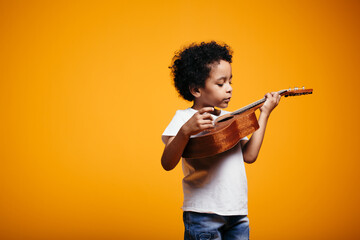 Boy in a white T-shirt holds a ukulele guitar near his chest and stares intently at strings against...