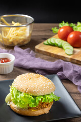 Crispy fried chicken burger on black plate served with french fries and ketchup on wooden background.
