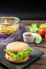 Crispy fried chicken burger on black plate served with french fries and ketchup on wooden background.