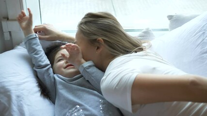 Young mom kisses daughter lying on bed near window, little girl goes slow motion