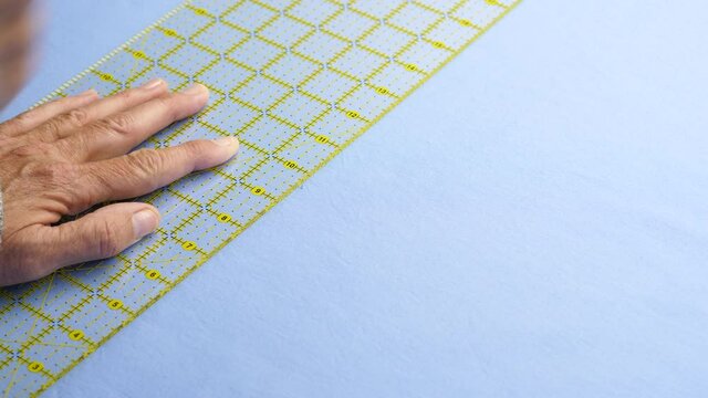 Female cuts blue cotton fabric using a yellow ruler to guide a rotary cutter on a green cutting mat 