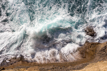 raging waves of turquoise color beat against the rocks on the beach and become white foam, brightly lit by the sun, view from above