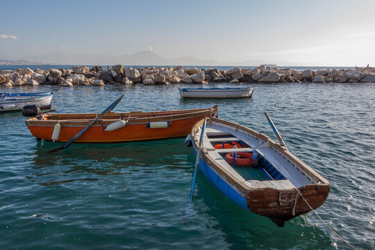 Fishing wooden boats with oars and motor in the turquoise water of the Bay of Naples against the background of a stone breakwater, in the haze of the mountains and the ship. Birds on the rocks.