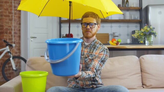 Man with bucket under umbrella suffering from leaking at home