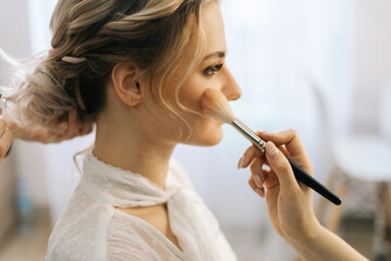 Fototapeta premium Close-up of make-up artist putting rouge on woman's cheek using brush while hairdresser making hair-do for young woman in beauty salon. Concept of backstage work.