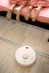 A young couple is resting in bed, and a robot vacuum cleaner cleans the wooden floor, modern...