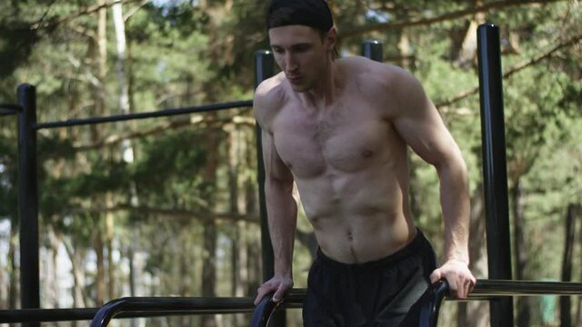 Adult caucasian handsome ath;ete is doing push ups on bars topless at the gym at the park during his workout
