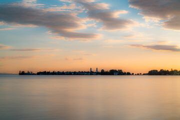 softer Blick auf die Skyline der Insel Lindau im Bodensee, im Abendlicht; Sonnenuntergang über der Insel Lindau im Bodensee mit orangeroter Sonne und blauem Himmel, glatte Wasserfläche