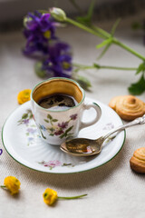 Espresso coffee in small delicate vintage cup with floral print, spoon, homemade cookies and spring flowers on linen background.