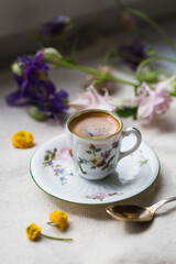 Espresso coffee in small delicate vintage cup with floral print, spoon and spring flowers on linen background.