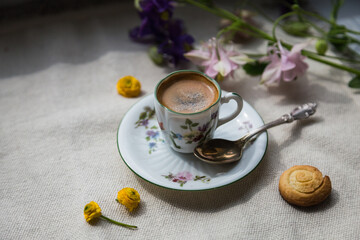 Espresso coffee in small delicate vintage cup with floral print, spoon and spring flowers on linen background.