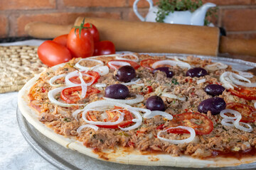 .Pizza of Brazilian cuisine with tuna stuffing with onion and olives, blurred background of tomatoes and wooden cylinder and bricks
