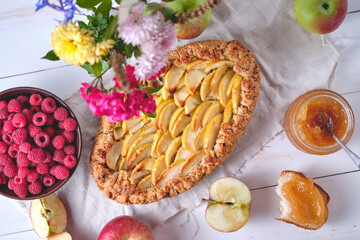 Homemade apple pie on a blackboard. Dessert on a white wooden table, around are apples, raspberries in a plate, apple jam and flowers. Top view.