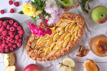 Homemade apple pie on a blackboard. Dessert on a white wooden table, around are apples, raspberries in a plate, apple jam and flowers. Top view.