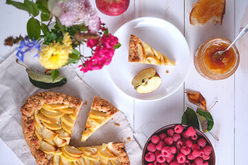 A sliced rustic apple pie on a linen towel. Dessert on a white wooden table, around are apples, raspberries in a plate, apple jam and flowers. Top view.