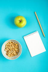 A cup with granola, a green apple and a notebook with a pencil to write calories on a blue background.