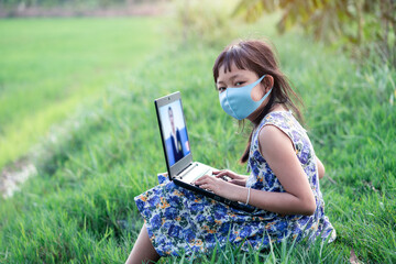 Happy little child girl learning outdoor by studying online and working on laptop in green field with wearing face mask